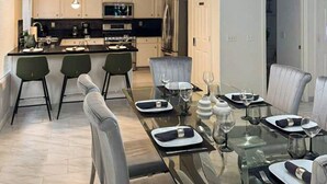 A modern kitchen with white cabinets, a central island, and a dining area with a glass table set for four. - 3BR/3BA w/ Pool & BBQ • 15 Mins to Strip & Airport (Las Vegas)