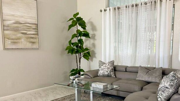 A modern living room with a glass coffee table, a grey sofa, a large window with white curtains, and a potted plant. - 3BR/3BA w/ Pool & BBQ • 15 Mins to Strip & Airport (Las Vegas)