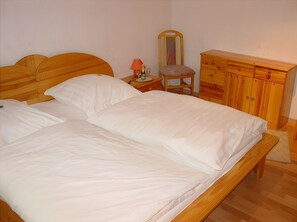 A wooden bed with white linens, a nightstand with a lamp, a chair, and a wooden dresser with drawers. - Holiday Home 'Seestern' with Private Terrace, Shared Garden and Wi-Fi (Wangerland)