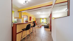 A kitchen with a counter, two bar stools, a dining area with a sofa and a coffee table, and a stone wall.