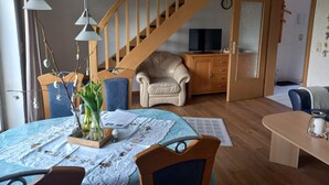 Olohuoneessa on puinen portaikko, televisio, sinisellä pöytäliinalla katettu ruokapöytä ja beigenvärinen nojatuoli.