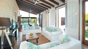 Ein großzügiger Wohnbereich mit einem weißen Sofa, einem Couchtisch und einem Fernseher. Durch die Glastüren ist ein Pool zu sehen.