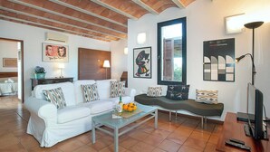 Ein Wohnzimmer mit einem weißen Sofa, einem Glastisch und einem dunklen Sofa. An den Wänden befinden sich Wandlampen, ein Fernseher und Kunstwerke.