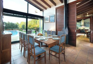 A dining area with a view of a swimming pool, wooden furniture, and a kitchen in the background.