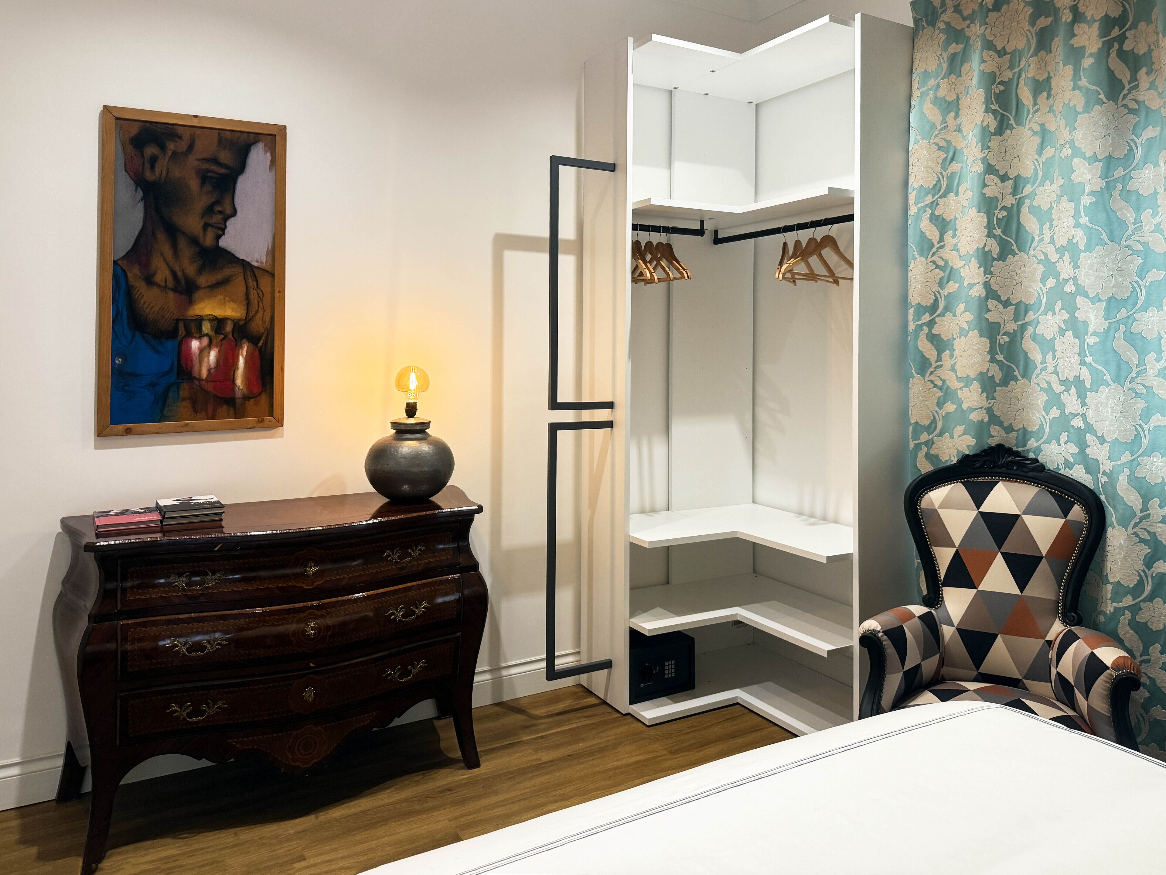 A bedroom with a wooden dresser, a painting, a bedside lamp, a wardrobe, and a patterned armchair.