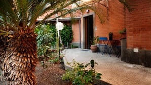 A courtyard with a palm tree, potted plants, a blue folding chair, and a brick building with an open door. - THE HOUSE OF JANUS (Ciampino)