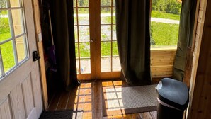 A wooden cabin interior with a door leading to a grassy area, a bench, and a trash bin. - Rustic tiny cabin-WiFi & power, close to beach, boat launch, lighthouse, trails (Rapid River)