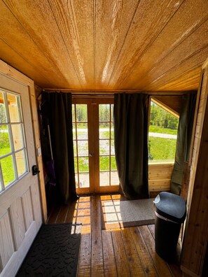 A wooden cabin interior with a door leading to a grassy area, a bench, and a trash bin. - Rustic tiny cabin-WiFi & power, close to beach, boat launch, lighthouse, trails (Rapid River)