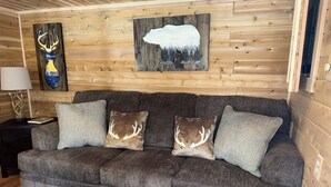 Interior de una cabaña de madera con un sofá gris, dos almohadones con motivos de ciervo, una lámpara y un cuadro con un ciervo y un oso.