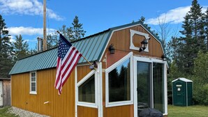 Un cobertizo de madera con un ventanal grande, una bandera estadounidense y un baño exterior verde.