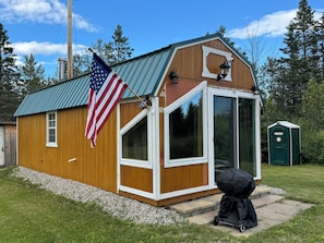 Un abri en bois avec une grande fenêtre, un drapeau américain et des toilettes extérieures vertes.