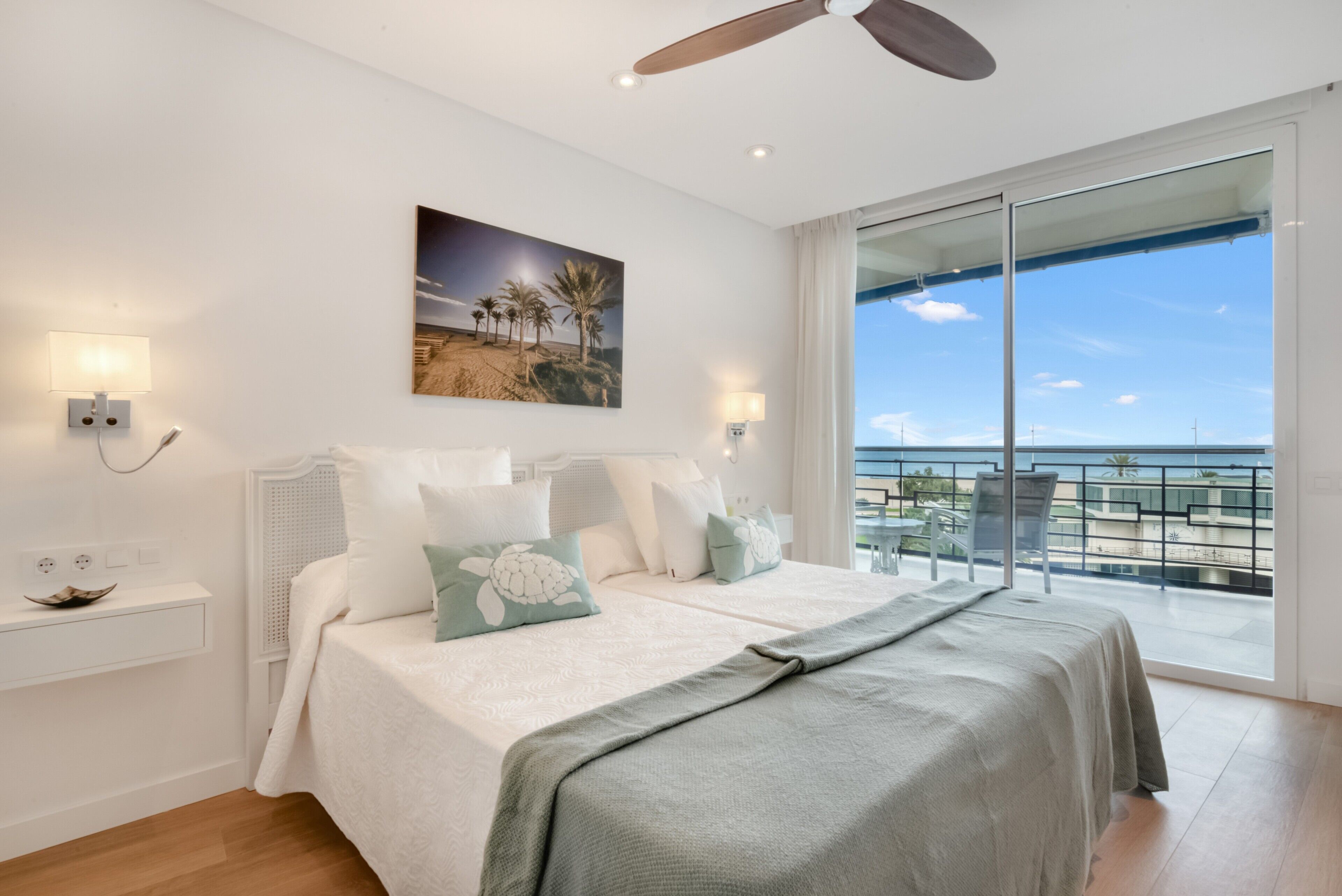 Ein Schlafzimmer mit Bett, Nachttisch, Wandlampe, Gemälde und einem Balkon mit Aussicht.