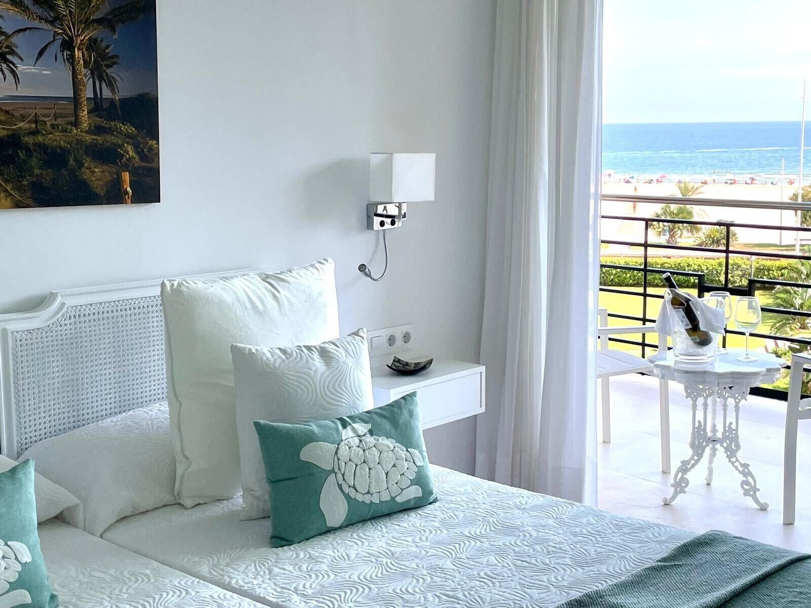 A hotel room with a bed, a nightstand, a wall-mounted phone, and a balcony with a view of the beach.
