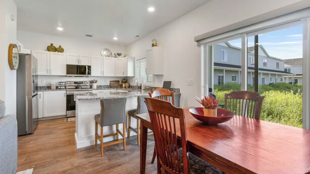 A modern kitchen with white cabinets, a central island, and a dining area with a wooden table and chairs. - 3BR, 2.5BA Townhome - Great Location + Open-Layout + Modern (Rexburg)