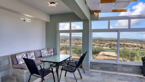 A sunlit patio with a table and chairs, a cushioned bench, and large windows offering a view of a hilly landscape. - Theasis Serenity Residence, Secluded Private Pool Retreat with Sea View (Sitia)