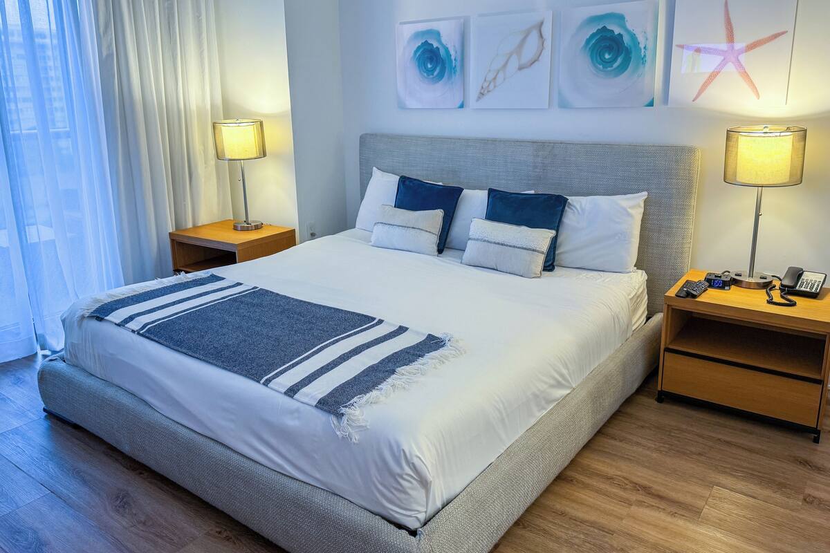 A neatly made bed with a blue and white striped throw, two bedside tables with lamps, and artwork on the wall.