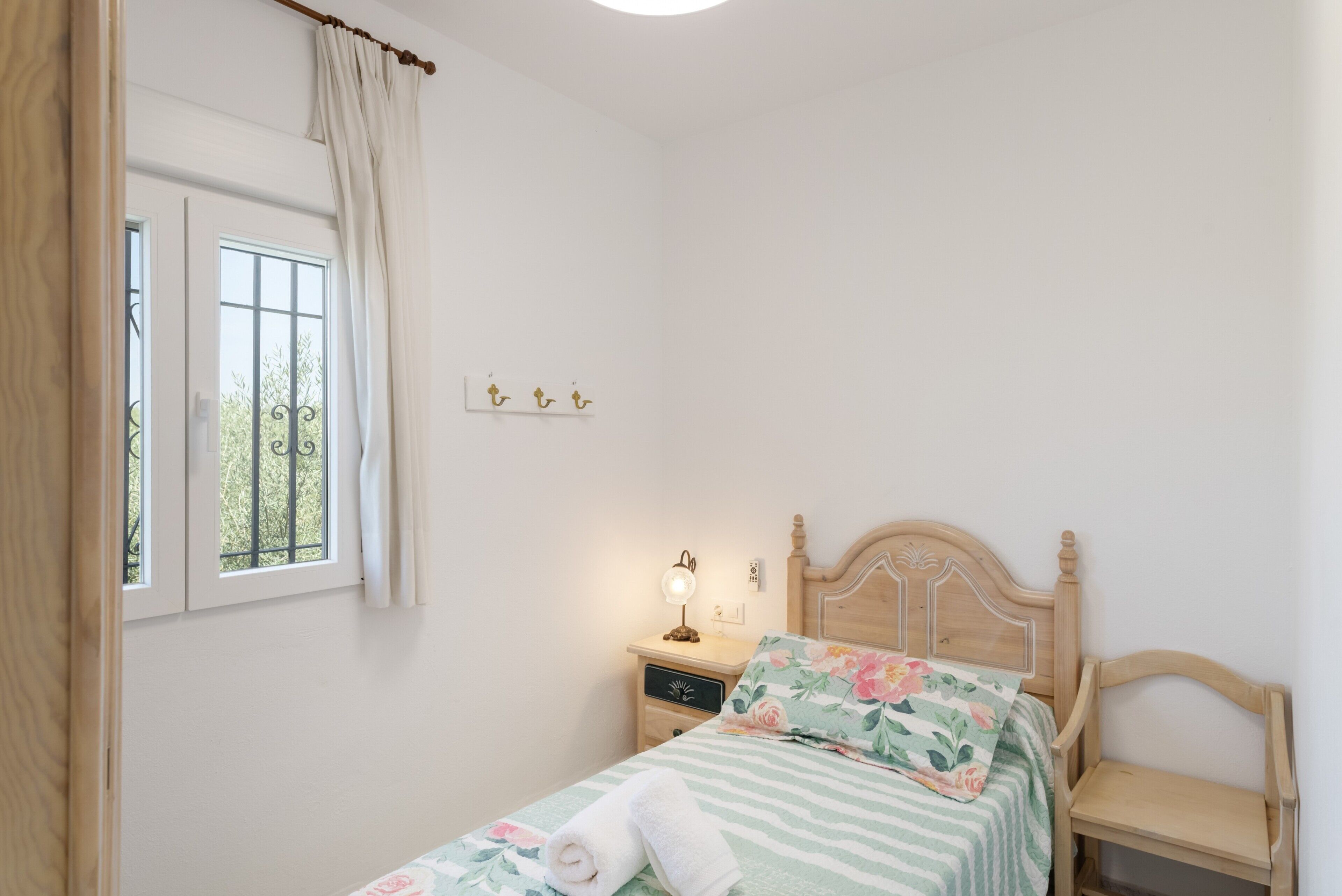 Un dormitorio con cama, mesita de noche, lámpara, ventana con cortinas y estante de pared con adornos.