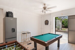 A pool table, foosball table, and bar stools in a room with a ceiling fan and a dartboard.