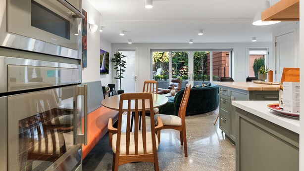 A modern kitchen with a dining area, featuring a built-in oven, a dining table with chairs, and a view of the outdoors through large windows.