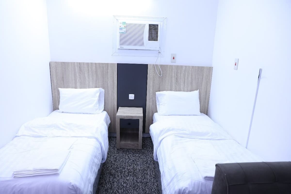 Une chambre d’hôtel avec deux lits simples, une petite table de chevet en bois, un climatiseur fixé au mur et une tête de lit de couleur foncée.