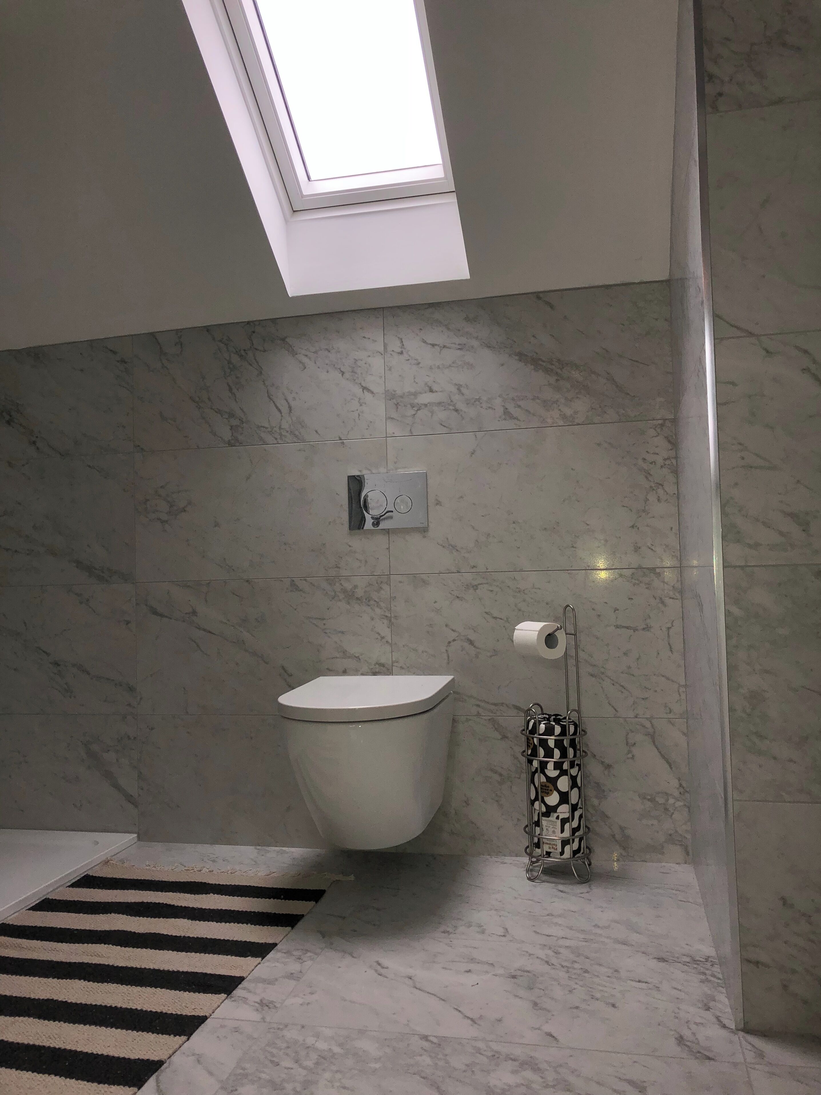 A bathroom with a skylight, a toilet, a marble floor, and a striped rug.