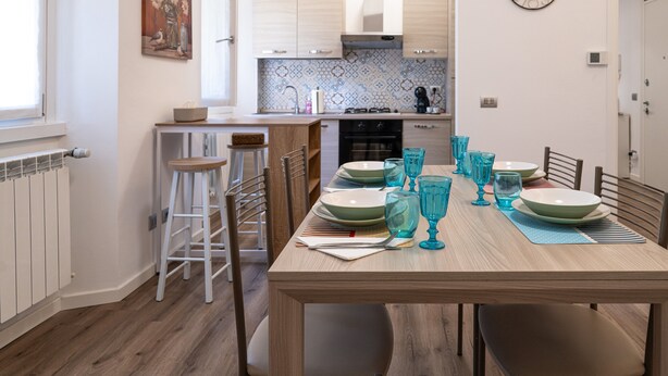 Un coin repas avec une table en bois dressée pour quatre personnes, des placards de cuisine, une horloge et un radiateur.