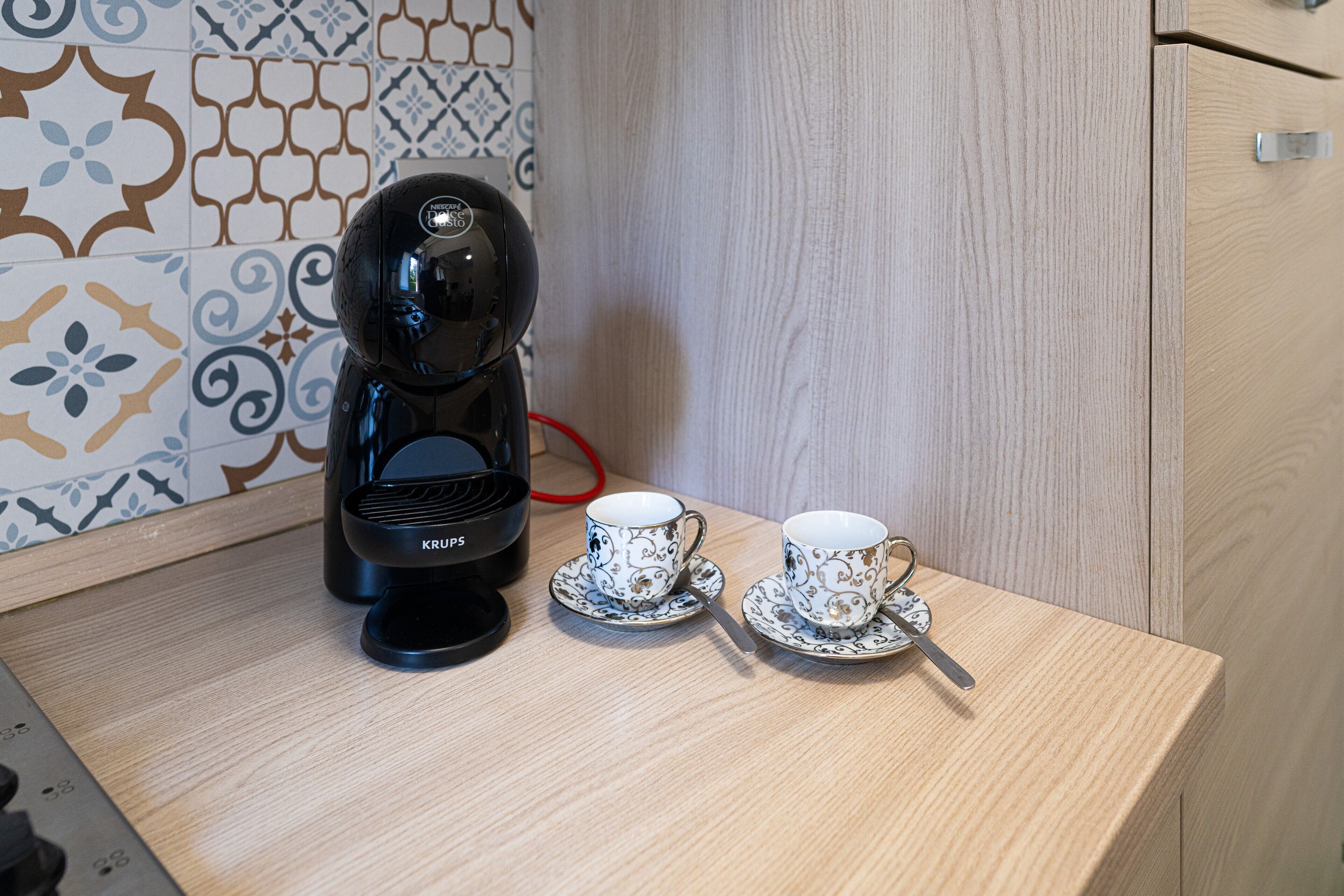 A coffee maker, two cups, and a spoon on a wooden table.
