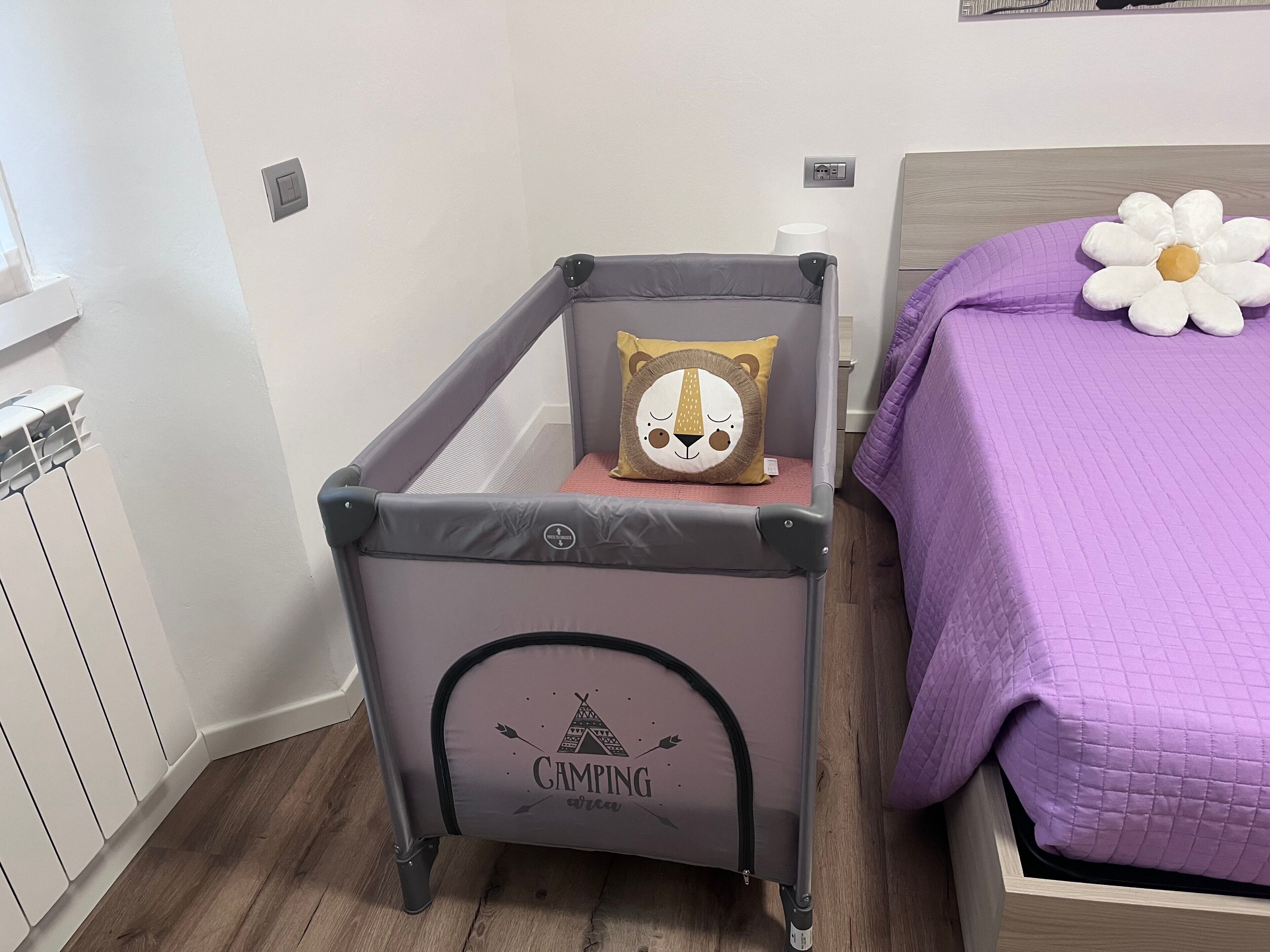 A room with a grey baby crib, a purple bed with a white flower pillow, and a wall with a power outlet.