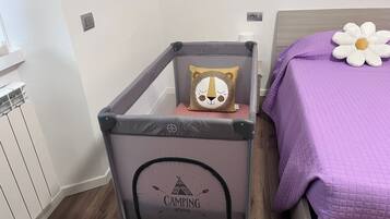A room with a grey baby crib, a purple bed with a white flower pillow, and a wall with a power outlet.