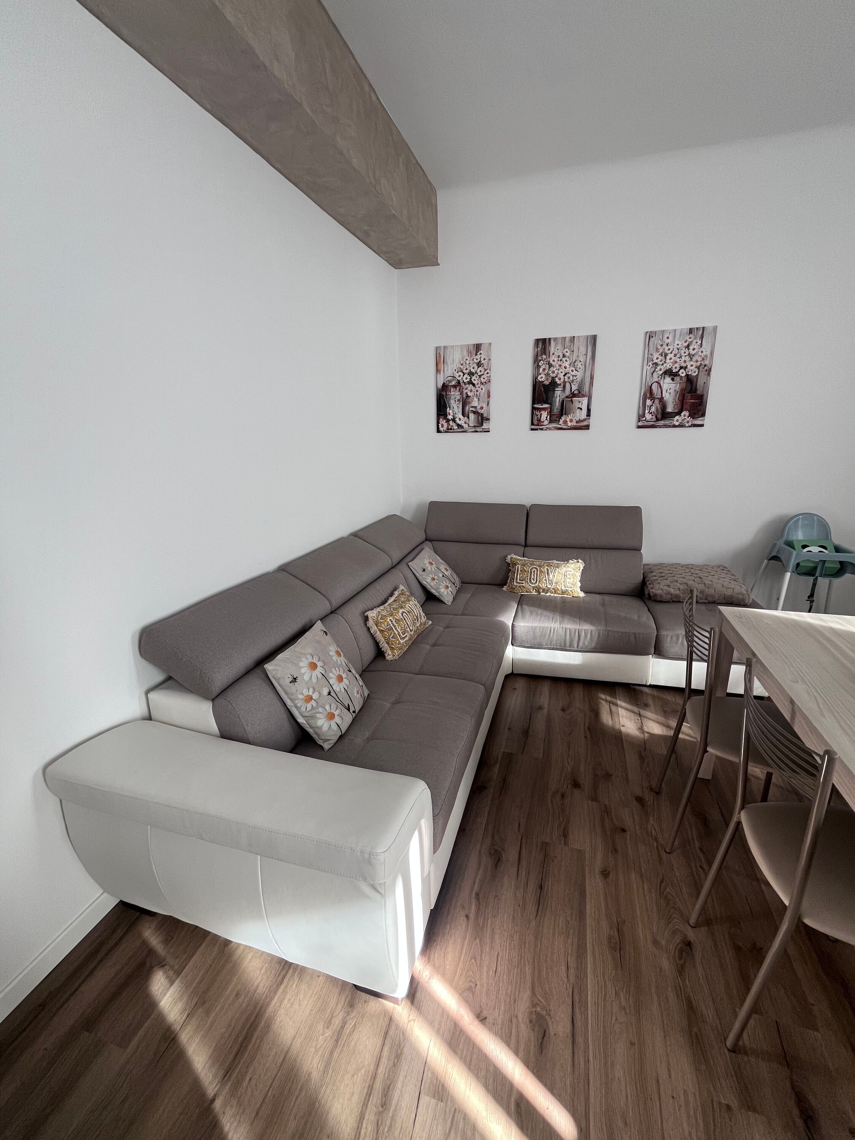 Un salon moderne avec un canapé d’angle gris, une petite table à manger et trois tableaux floraux accrochés au mur.