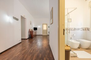Un corridoio con pavimento in legno, una televisione, una piccola scrivania e una porta che conduce a un bagno con vasca e servizi igienici.