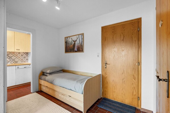 A small bedroom with a single bed, a wooden door, a small kitchenette, and a wall-mounted picture.