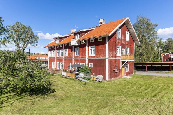 A red wooden multi-story building with a sloped roof, a covered porch, and a small garden area.