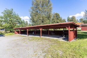 Katettu ulkoalue, jossa on istumapaikkoja. Sinne johtaa sorapolku, ja aluetta ympäröivät puut. Taustalla näkyy punainen rakennus.
