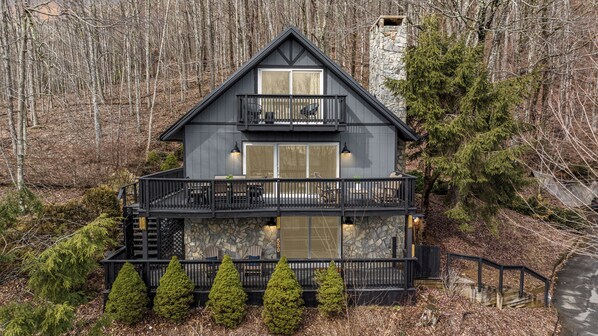 A two-story cabin with a balcony, stone facade, and a forest backdrop.