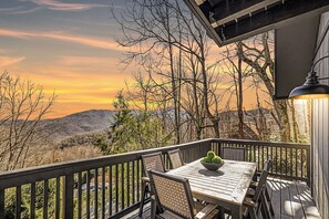 A wooden deck with a table and chairs, overlooking a mountainous landscape during sunset.