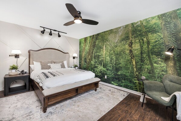 A bedroom with a large bed, a ceiling fan, a wall mural of a forest, a nightstand, a lamp, and a chair.