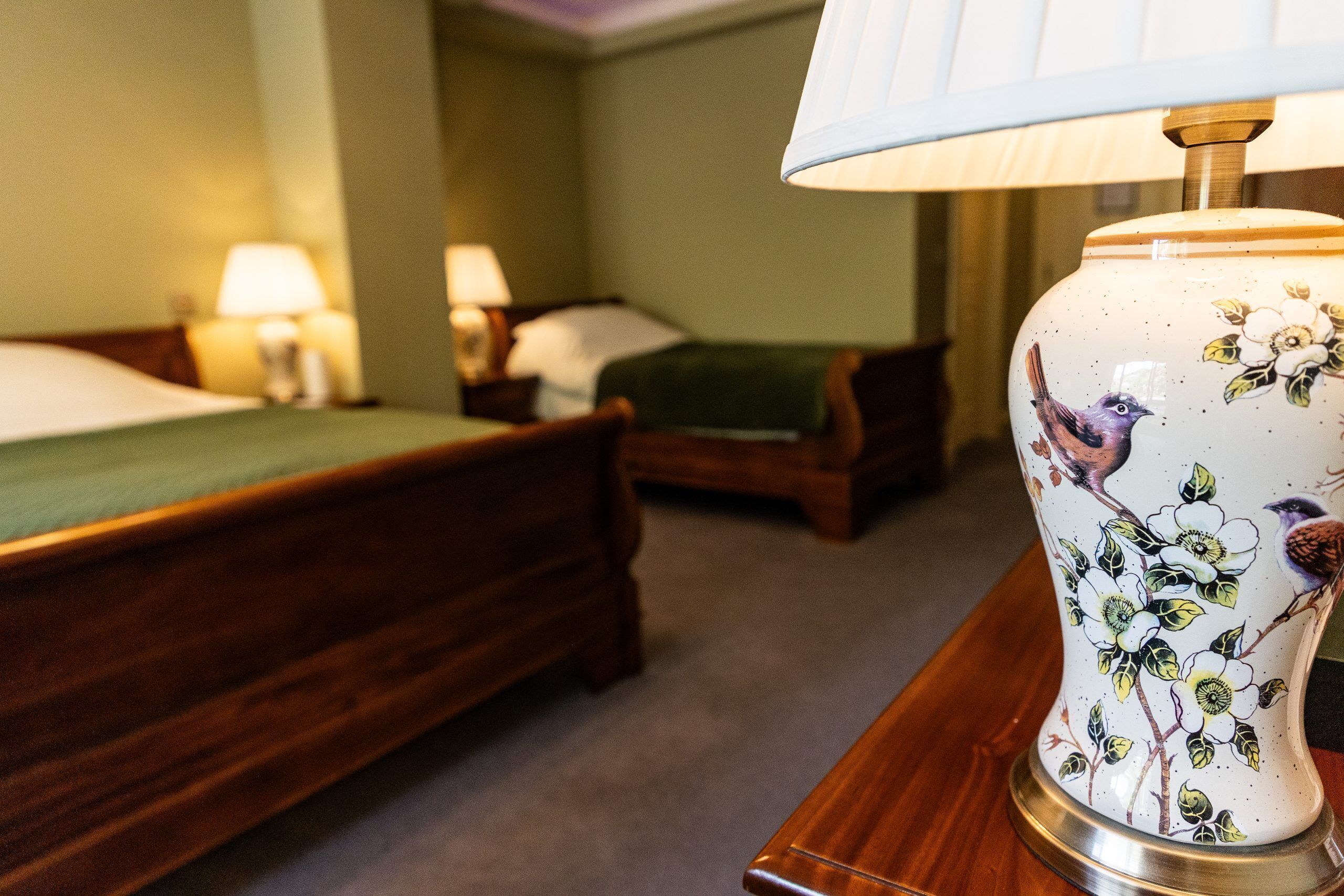 Ein Hotelzimmer mit zwei hölzernen Betten, einer grünen Decke auf einem Bett und einer dekorativen Vase mit floralen und Vogelmotiven auf einem Nachttisch.