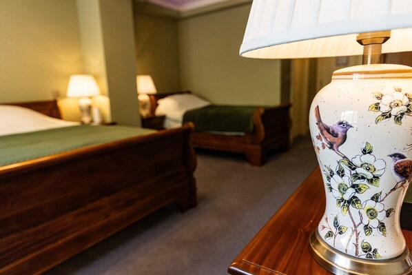 A hotel room with two wooden beds, a green throw on one bed, and a decorative vase with floral and bird designs on a nightstand.