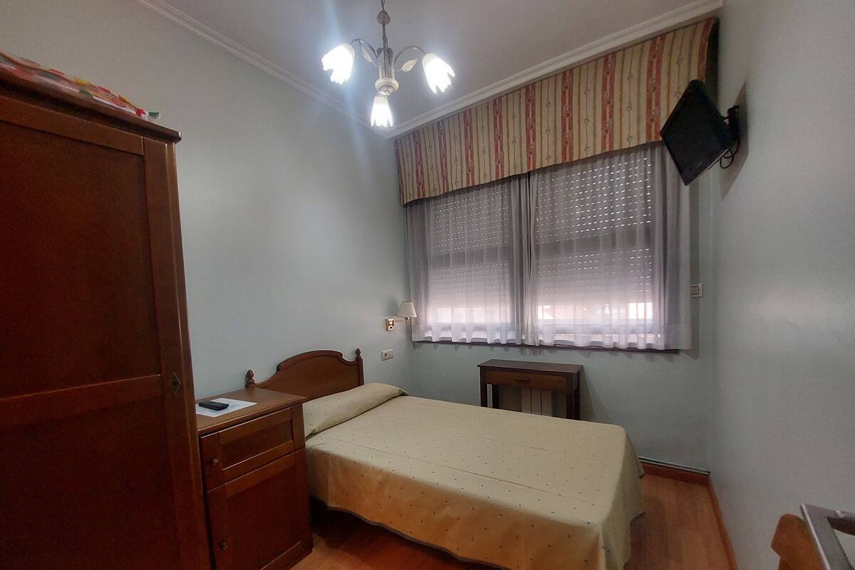 A bedroom with a wooden wardrobe, a single bed with a white bedspread, a bedside table with a lamp, and a window with striped curtains.