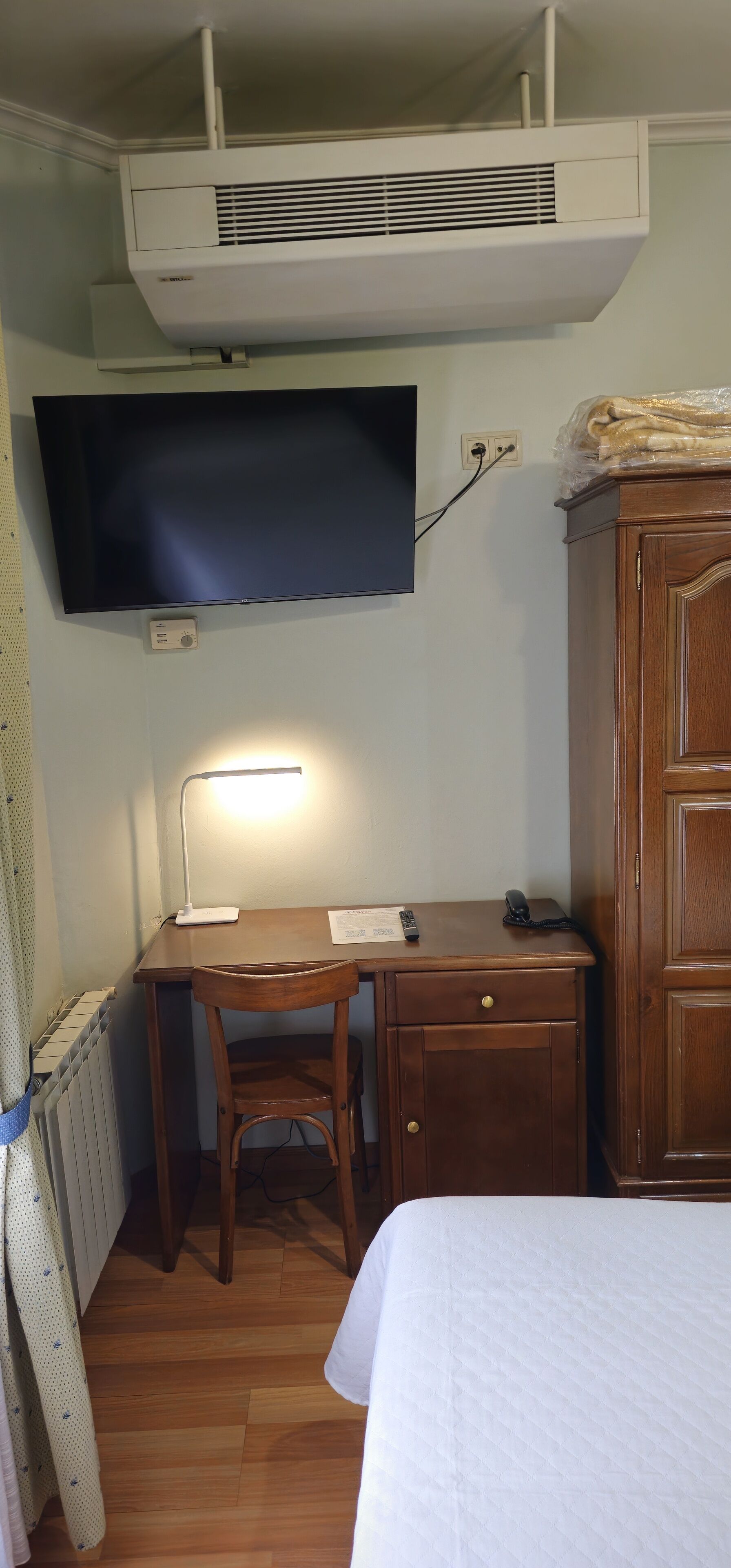 A room with a bed, a desk with a chair, a television mounted on the wall, and a radiator.