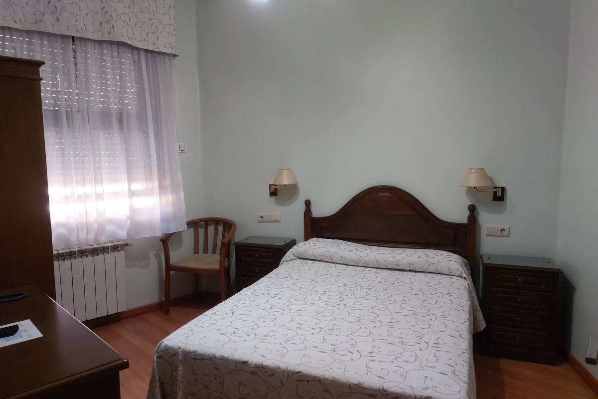 A bedroom with a bed, two bedside tables, a chair, a desk, and a radiator.