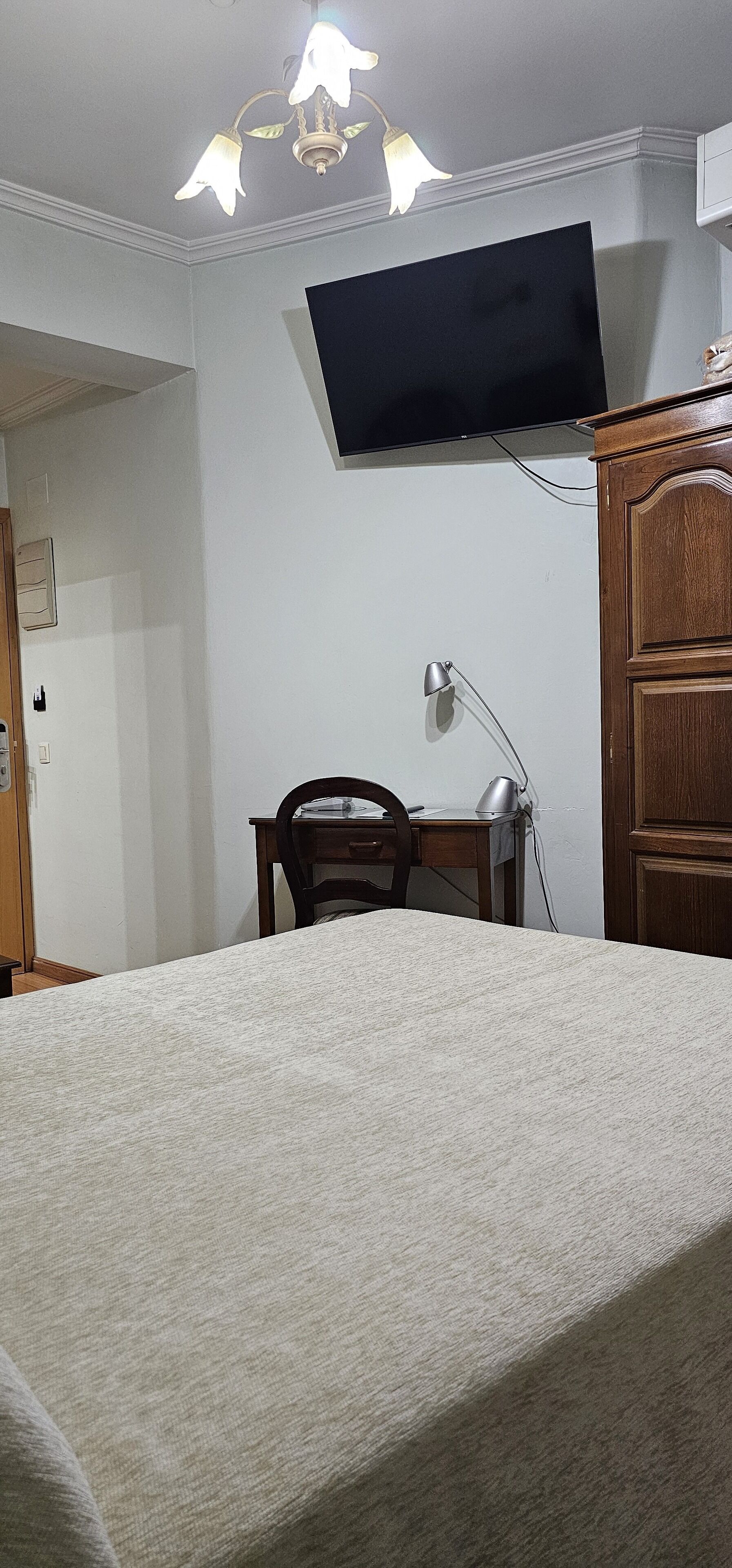 A bedroom with a bed, a desk with a chair, a desk lamp, a flat-screen TV mounted on the wall, and a wooden wardrobe.