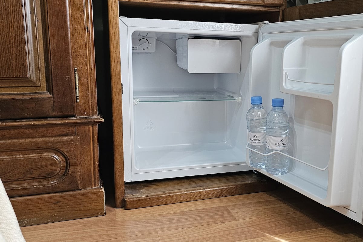 A small, white refrigerator with two bottles of water on the door shelf, located in a wooden cabinet with a wooden floor.