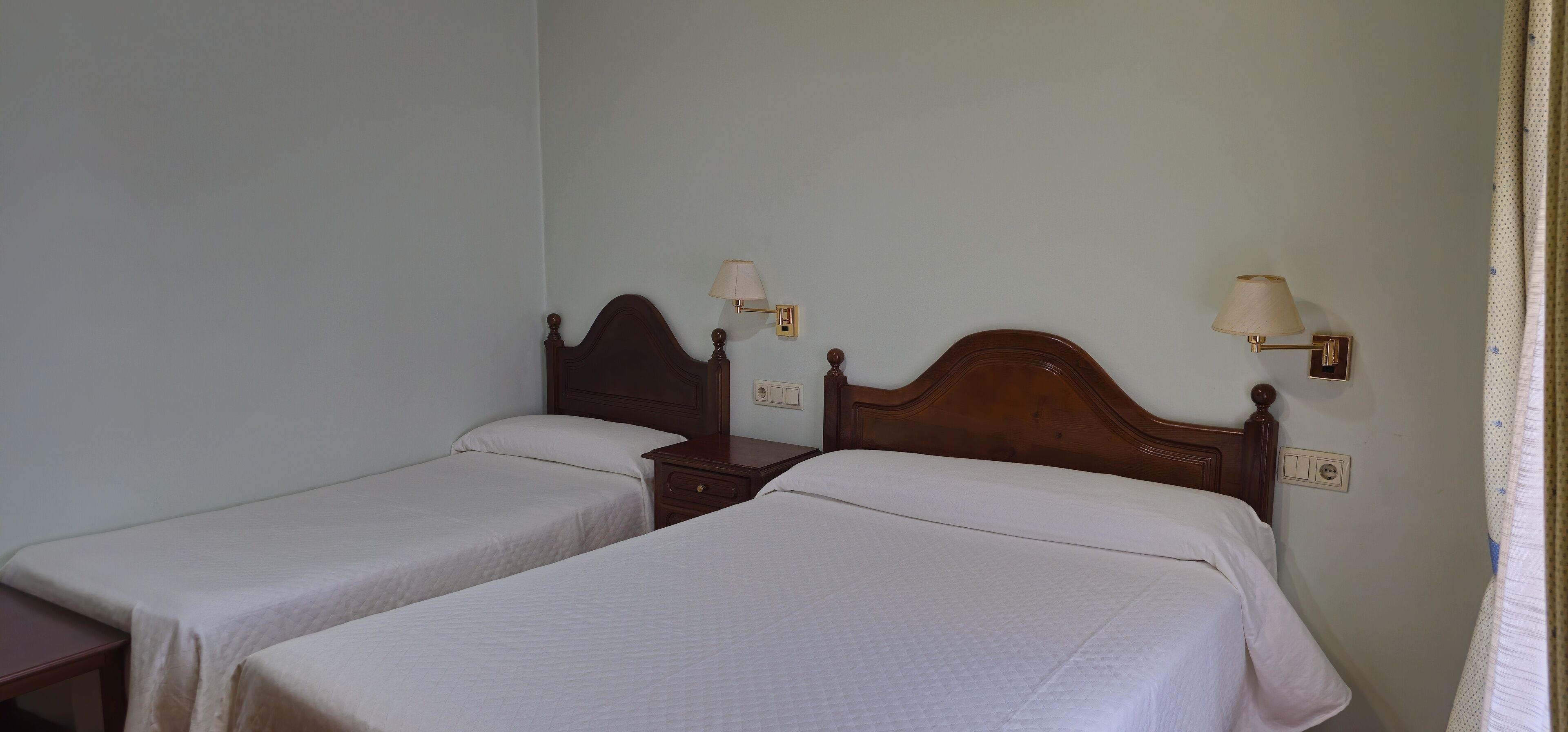 Una habitación con dos camas individuales, cada una con cabecera de madera y mesita de noche. Hay lámparas de pared a ambos lados de las camas.