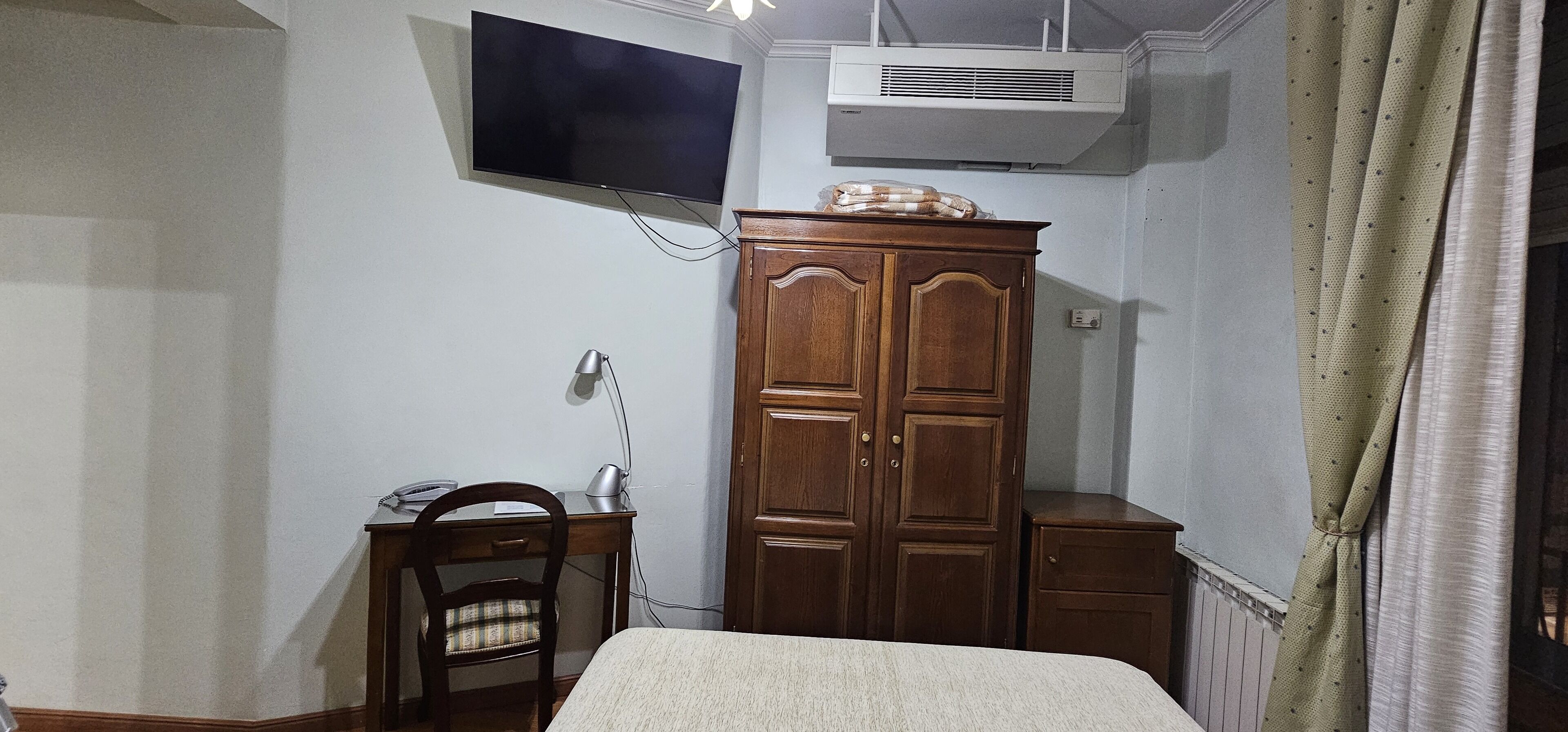 A bedroom with a bed, a wooden wardrobe, a desk with a chair, a flat-screen TV mounted on the wall, and an air conditioning unit.