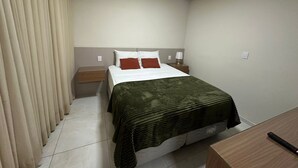 Una habitación de hotel con cama, mesita de noche, lámpara y ventana con cortinas.