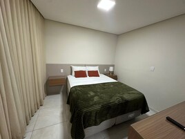 Quarto de hotel com cama, mesa de cabeceira, luminária e janela com cortinas.