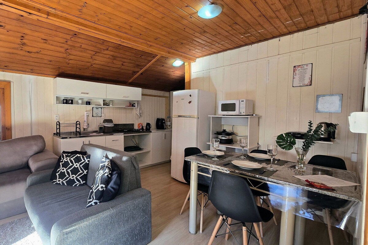 Cocina compacta con microondas, cafetera y refrigerador. Cuenta con una mesa de comedor con sillas, un sofá con cojines estampados y una ventana con vistas al exterior.