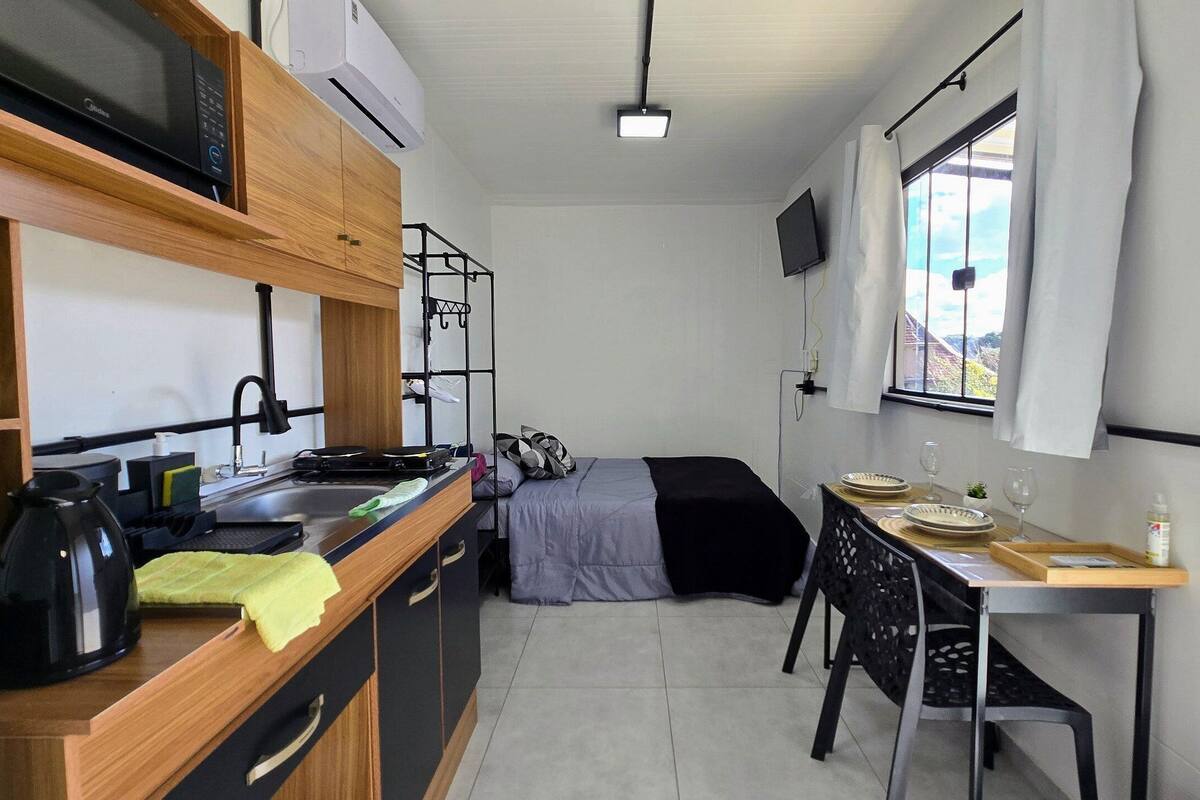Una kitchenette compacta con estufa, pileta y microondas, un pequeño comedor con mesa y sillas, y un ventanal con cortinas.